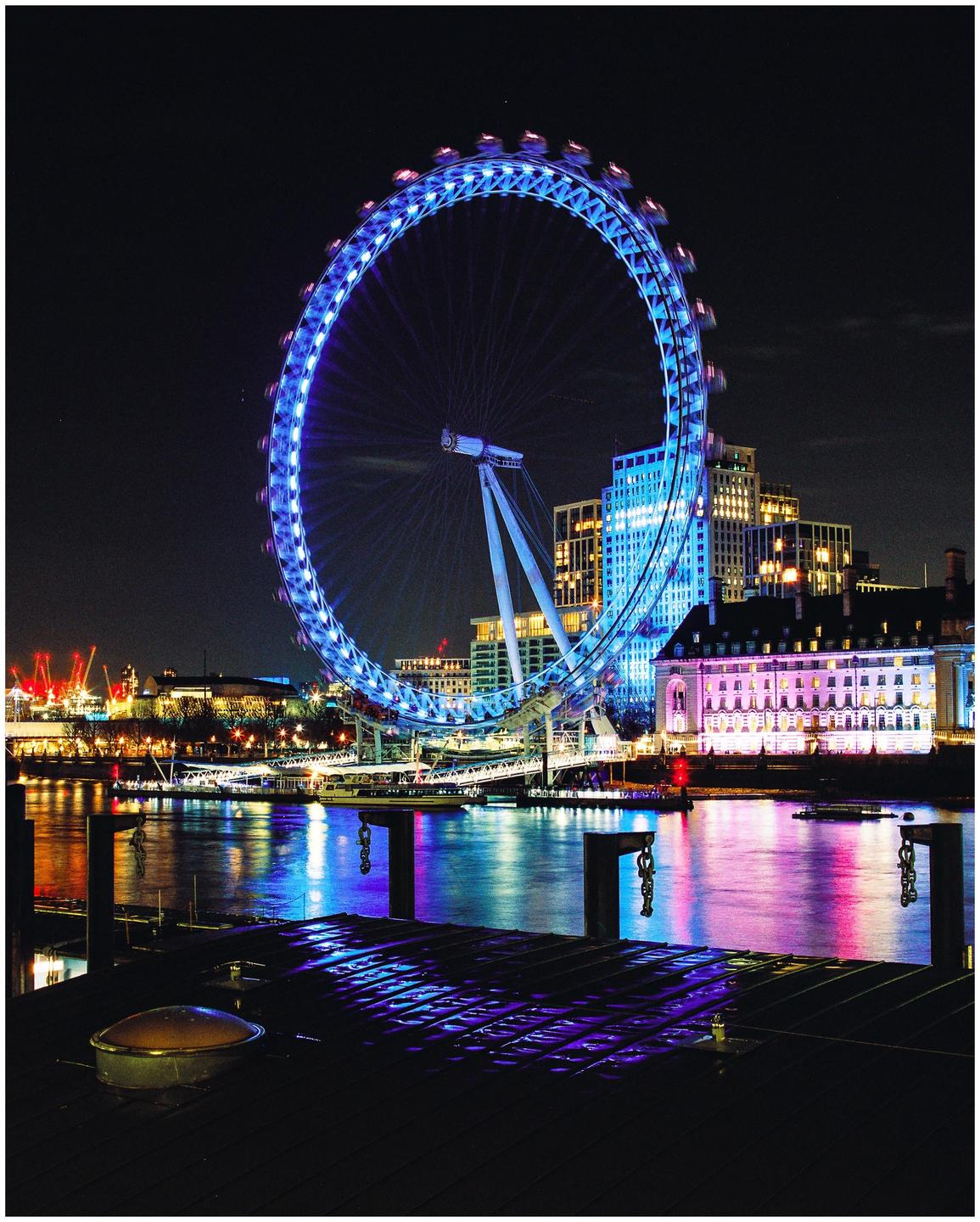 The London Eye is illuminated in blue at night, with its reflection visible in the water below.