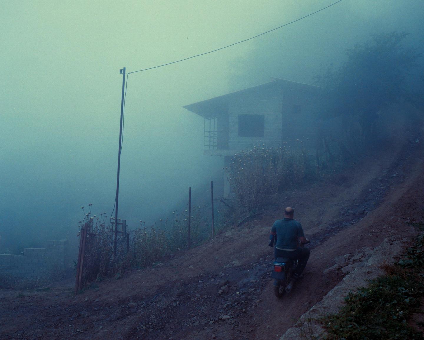 A man rides a motorcycle up a dirt path towards a house shrouded in fog.