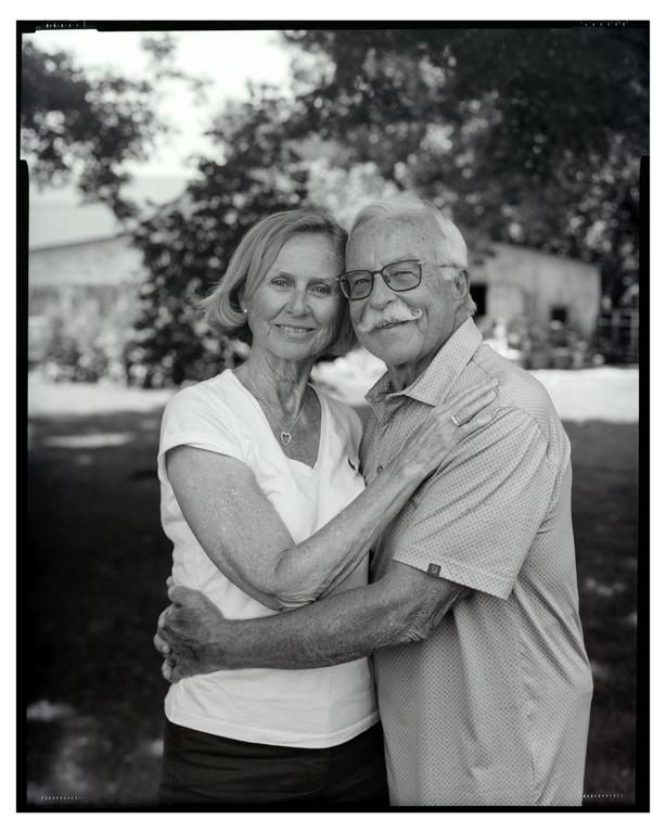 A man and woman embrace and smile at the viewer in a black and white outdoor portrait.