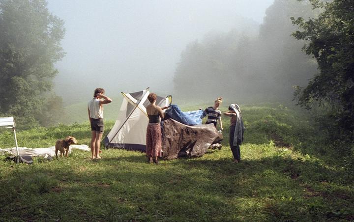Four people and a dog are gathered around a tent in a grassy, hazy field.