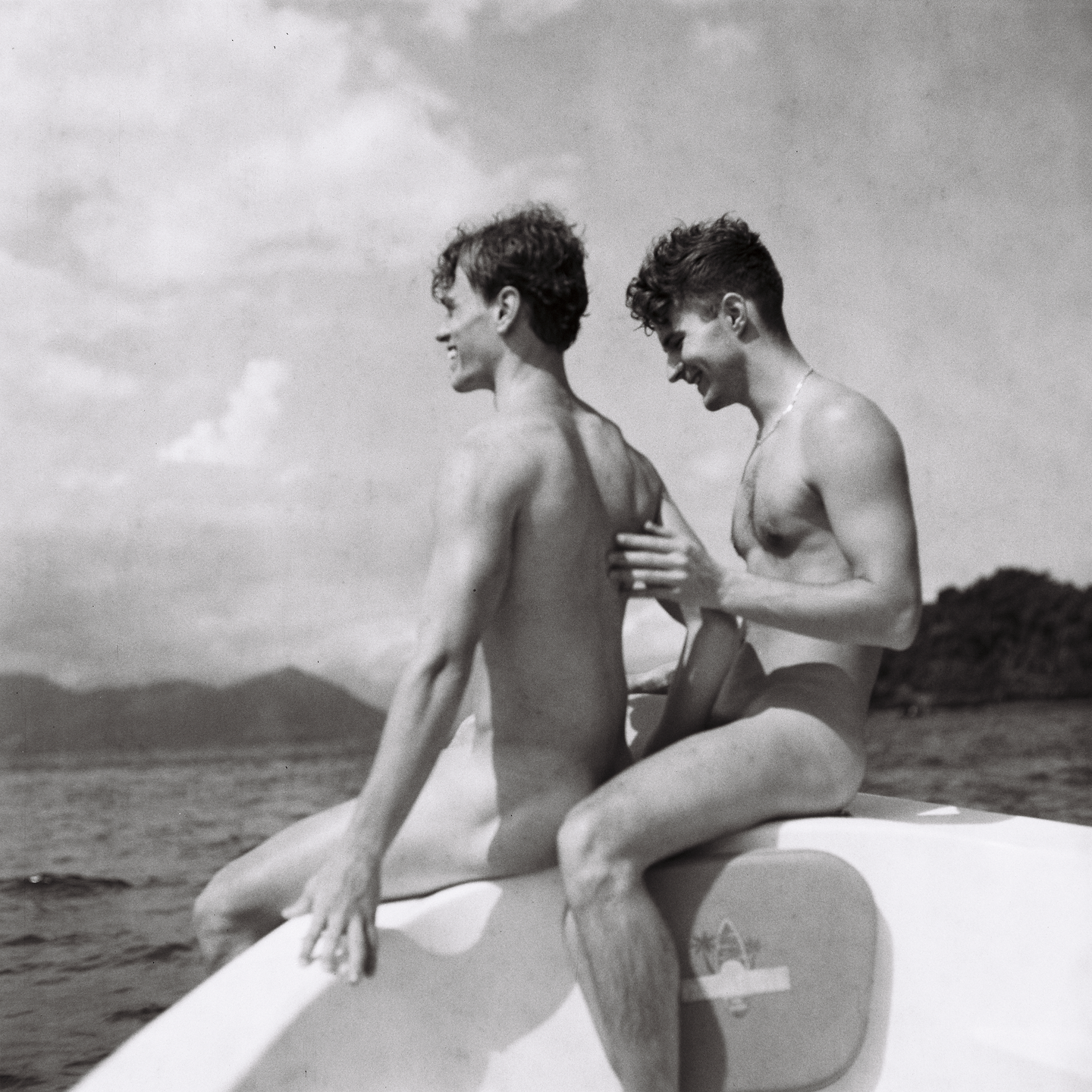 Two men sit on a boat on the water with mountains in the distance.