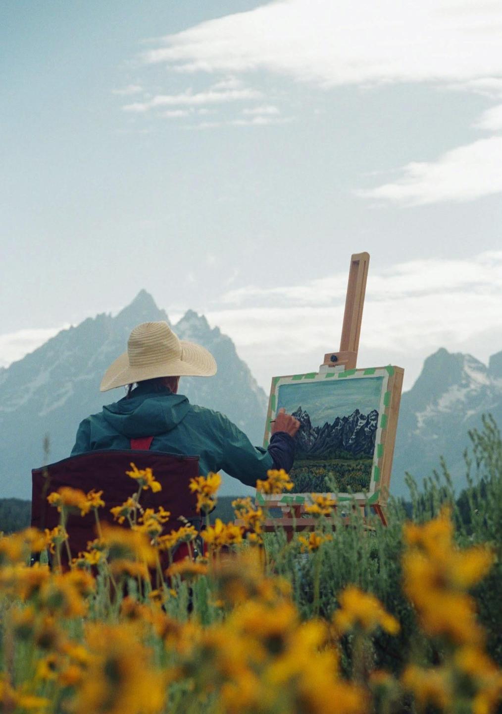 A person wearing a straw hat and a green jacket is painting a mountain landscape on an easel, surrounded by yellow flowers.