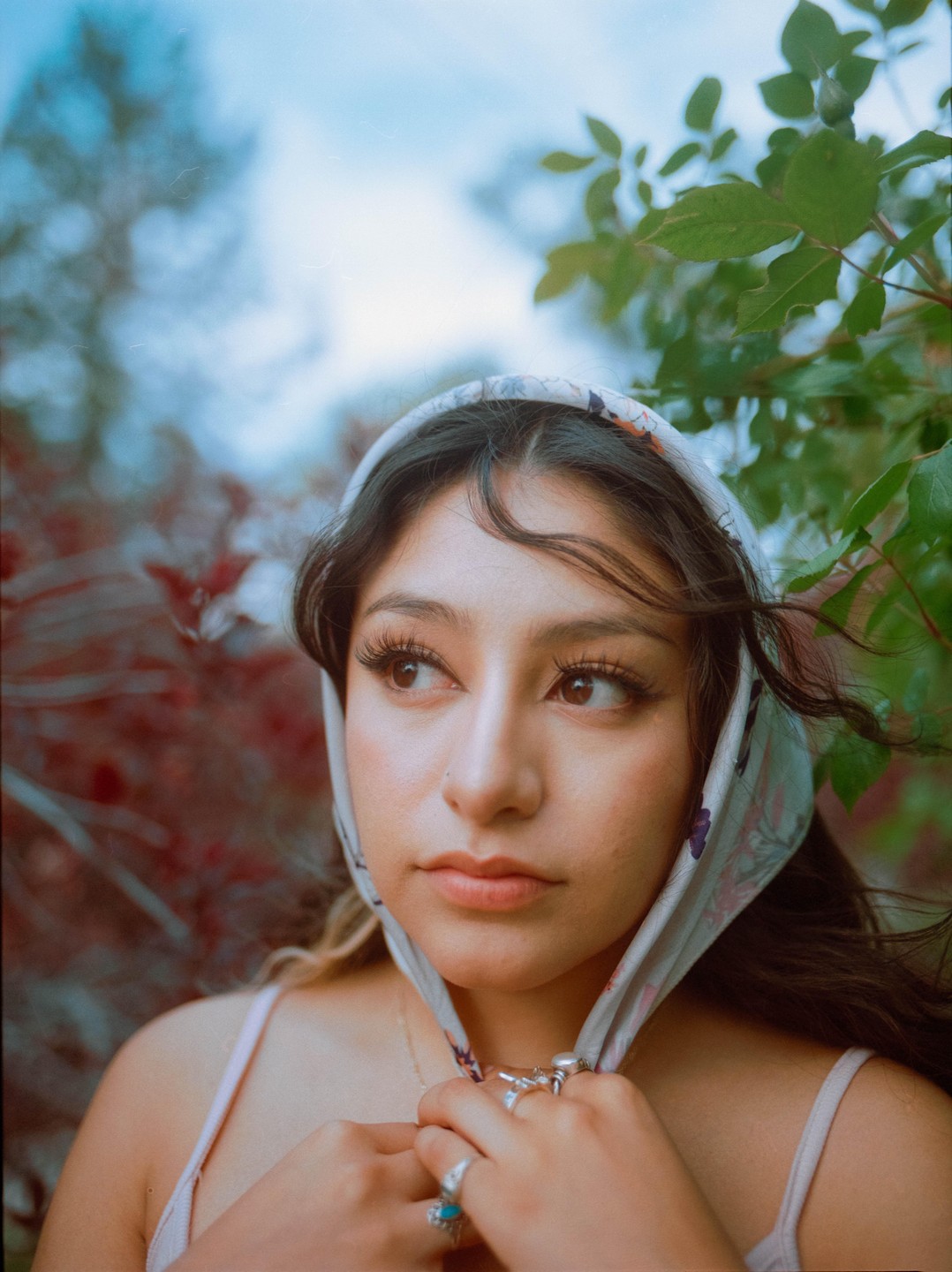 A woman wearing a headscarf looks to the side, surrounded by foliage.