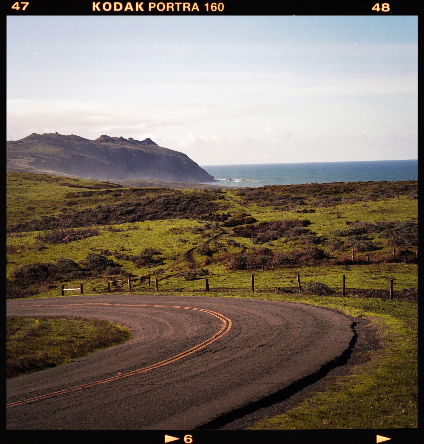 A winding road curves through a green, hilly landscape with the ocean and a distant mountain under a hazy sky.