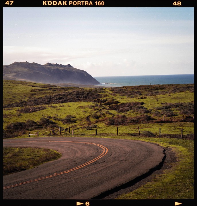 A winding road curves through a green, hilly landscape with the ocean and a distant mountain under a hazy sky.