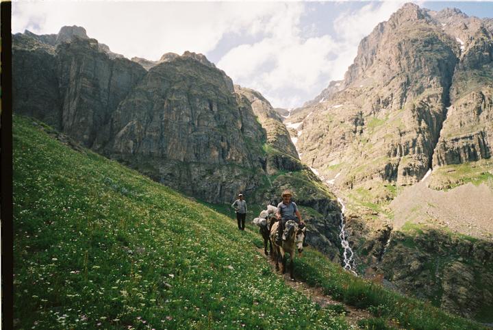 A man rides a horse with packed supplies while another man walks behind him on a narrow path through a mountainous landscape with a waterfall.