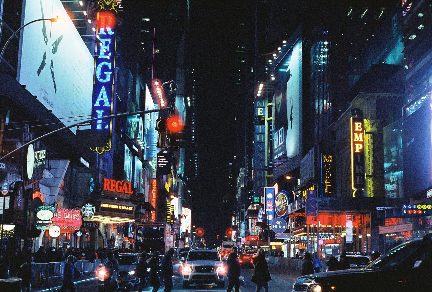 A street in New York City is bustling with people and cars at night, illuminated by numerous neon signs and billboards.