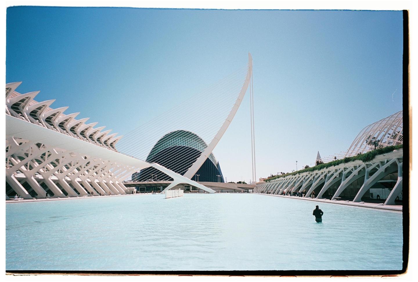 A person stands in a shallow pool of water surrounded by modern architecture under a clear sky.