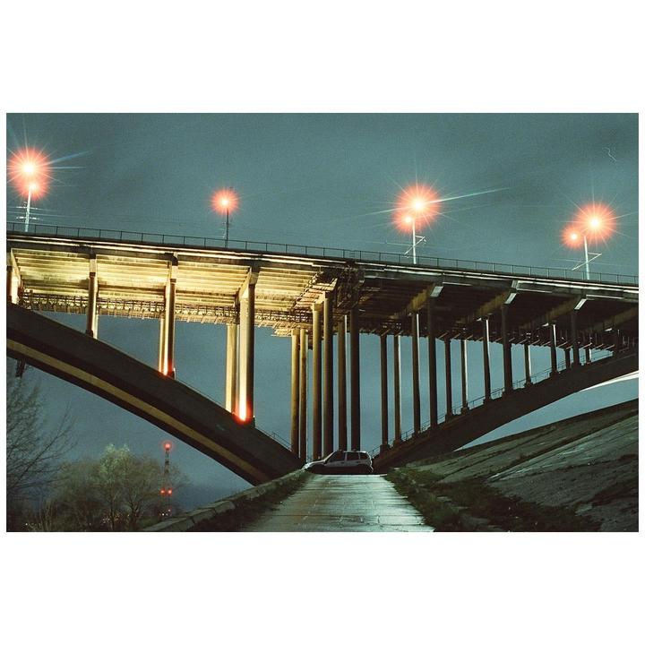 A car is parked on a wet road under a large bridge at night.