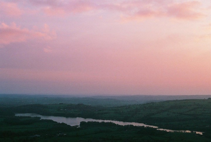 A wide shot shows a lake surrounded by green hills under a pastel pink and purple sky.