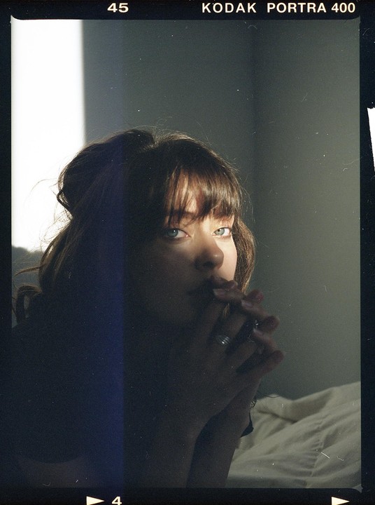 A woman with bangs and a ring on her finger looks at the camera, with light illuminating her face from the side.
