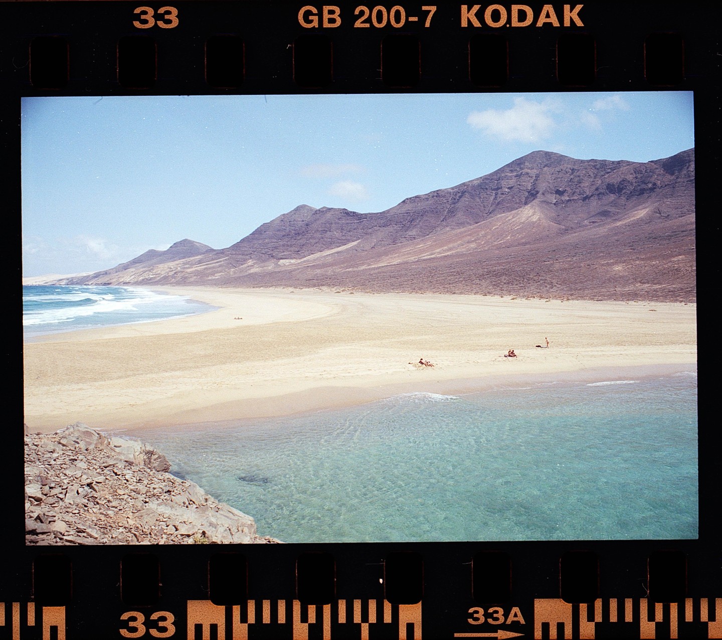 A wide sandy beach with clear turquoise water is backed by large mountains under a blue sky.