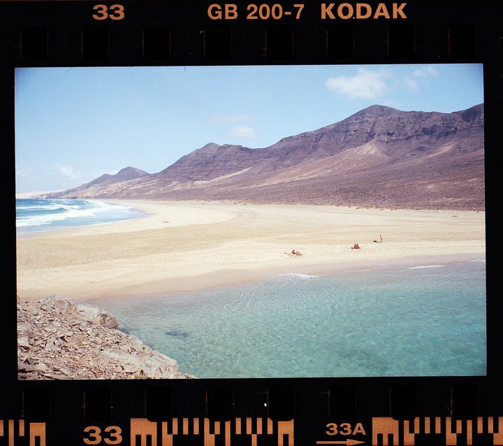 A wide sandy beach with clear turquoise water is backed by large mountains under a blue sky.