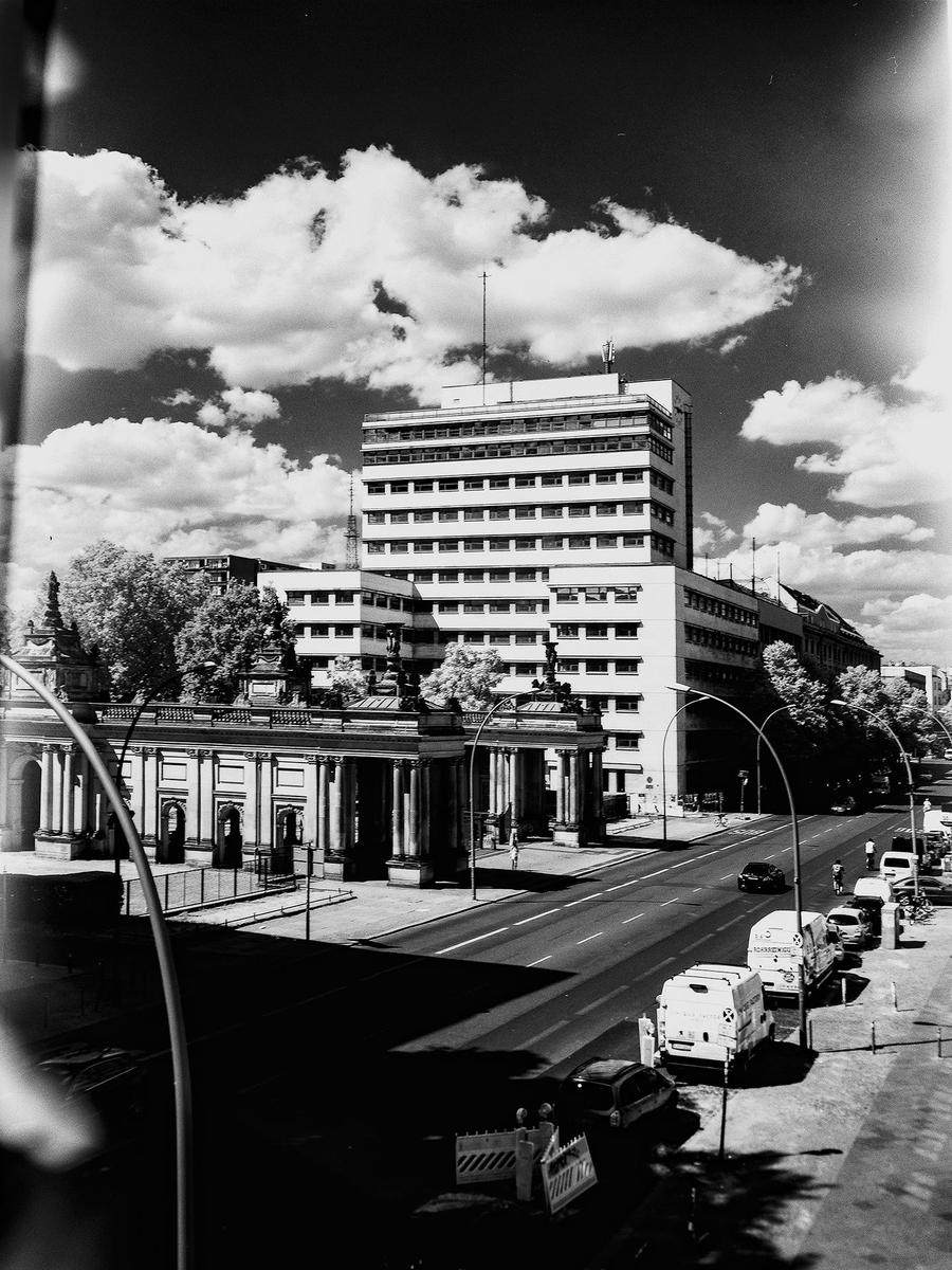 A tall building stands behind a classical colonnade next to a street with parked cars.