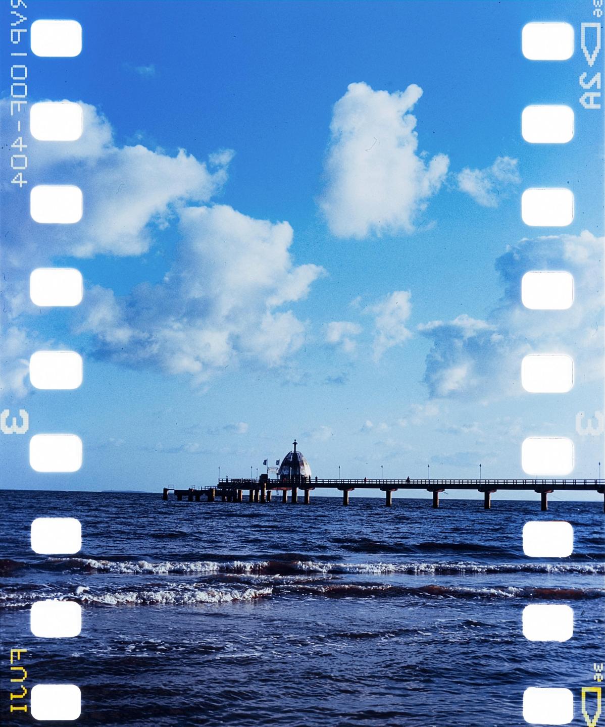A long pier with a domed building at its end extends into the ocean under a blue sky with white clouds.