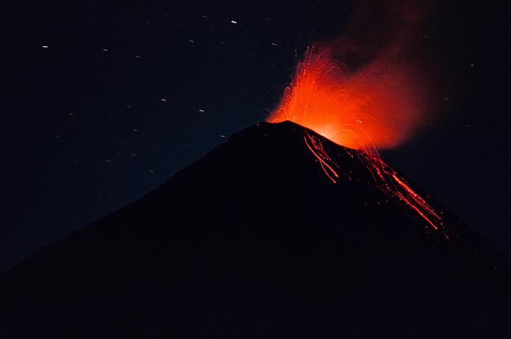 A volcano erupts at night, spewing bright orange lava and smoke into the dark sky.