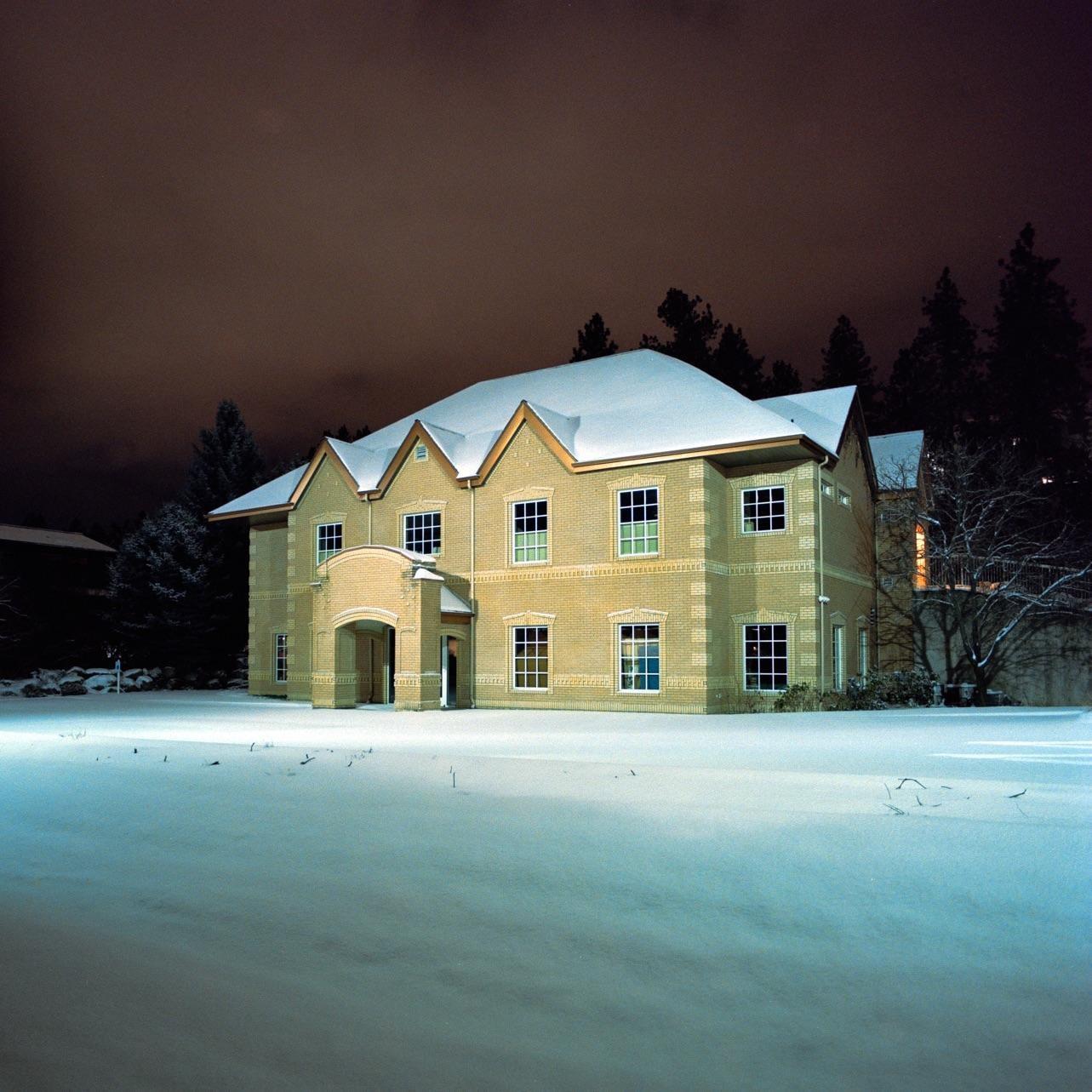 A large brick house with a snow-covered roof stands in a snowy field at night, with trees in the background.