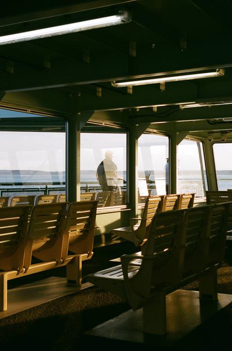 A man stands looking out the window of a ferry with rows of empty seats in the foreground.
