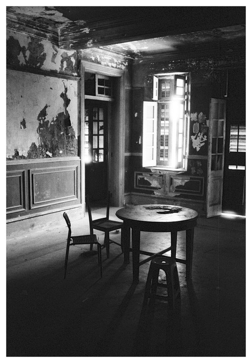 Sunlight streams through a window into an abandoned room with peeling paint, a round table, and three chairs.