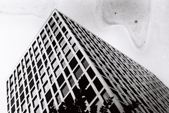 A tall building with a grid pattern of windows is seen from a low angle against a bright sky.