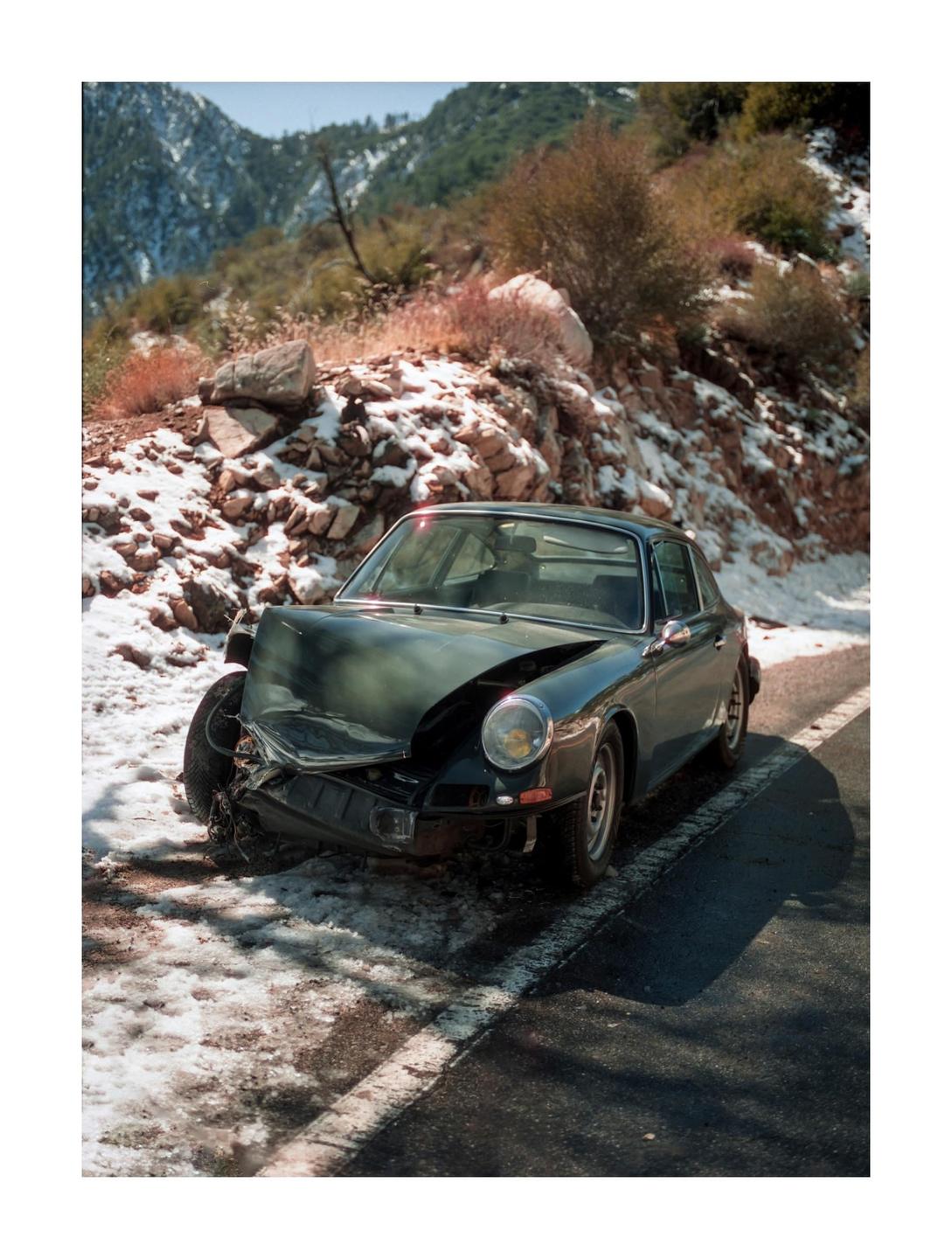 A dark green car with a damaged front end is parked on the side of a snowy mountain road.