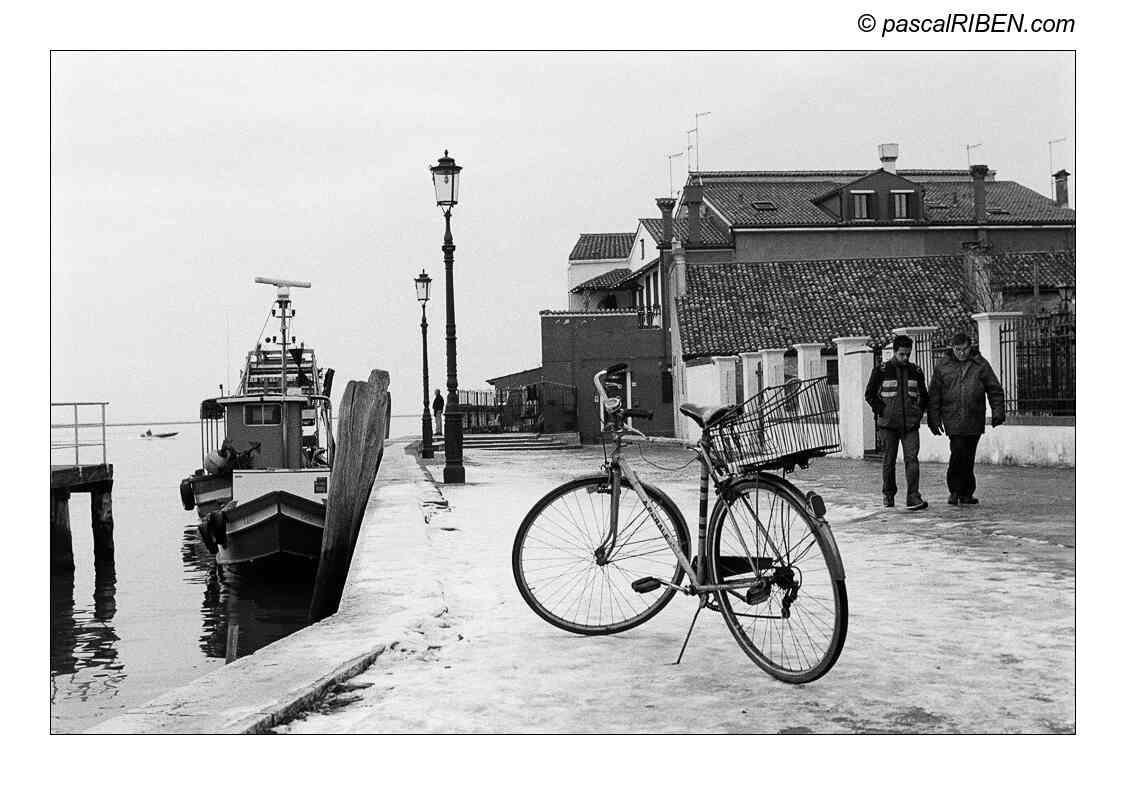 A bicycle with a basket is parked on a snowy dock next to a boat, with two men walking in the background and buildings across the water.
