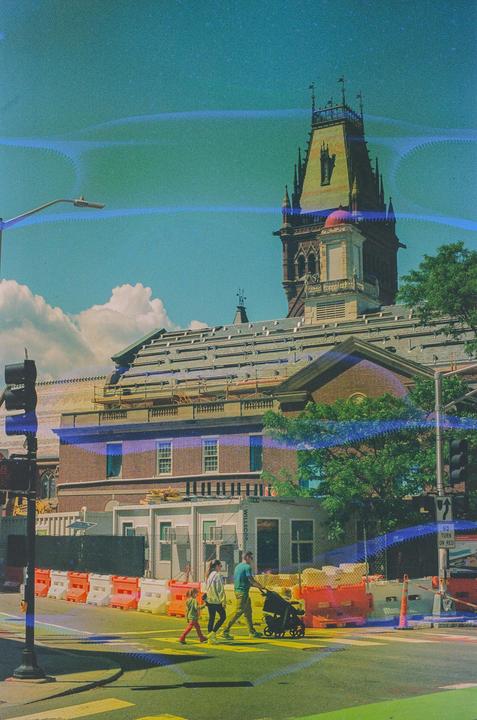 A family crosses a street in front of a large building with a prominent tower and scaffolding.