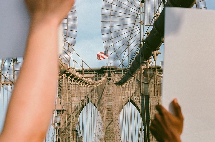 Two arms hold up white sheets of paper, framing the Brooklyn Bridge with an American flag flying at its top.