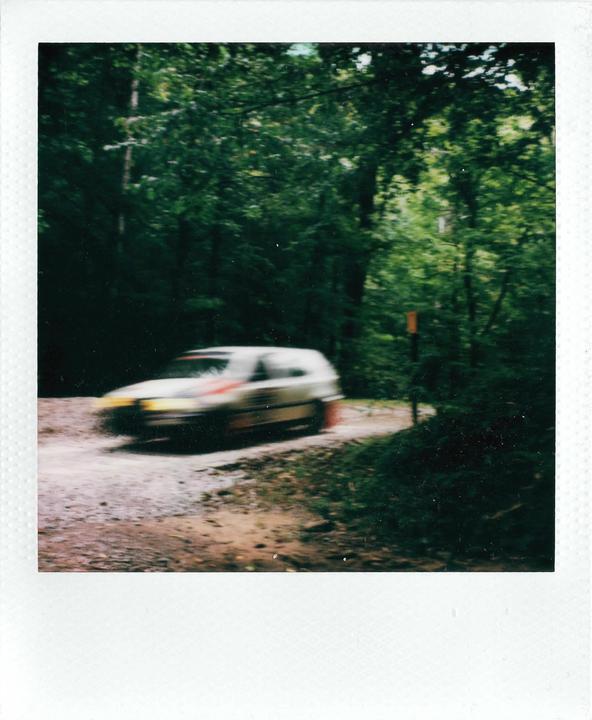 A blurry white rally car speeds along a dirt road through a forest.