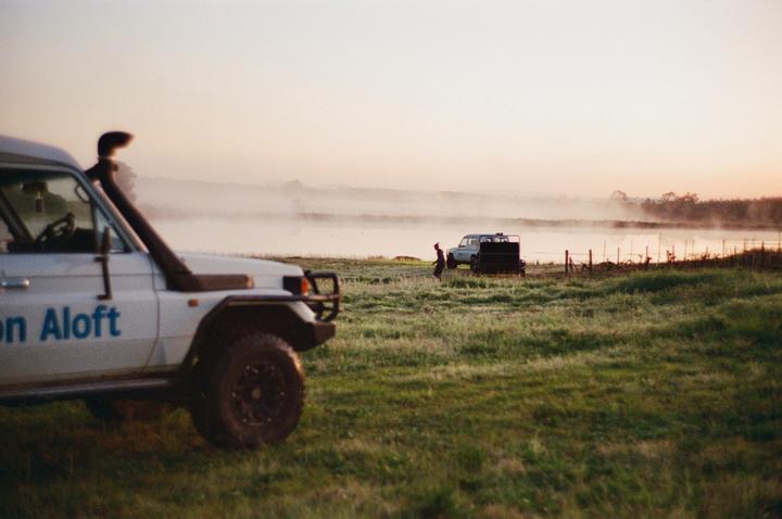 Two white vehicles are parked in a grassy field next to a body of water with fog rolling over it at dawn.