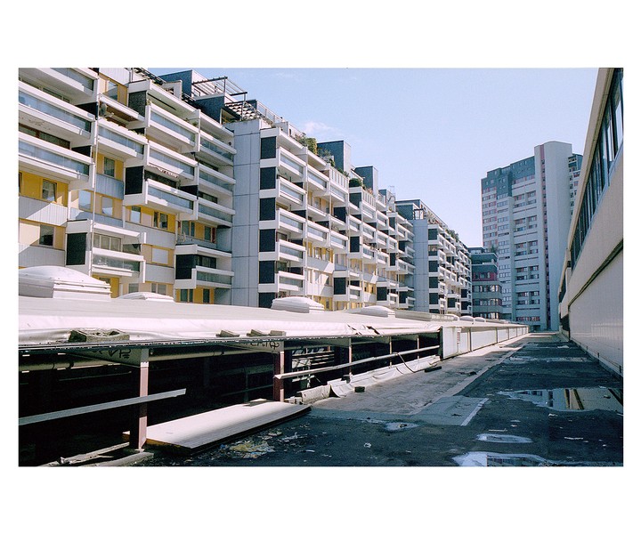 A rooftop with puddles and a long building with many balconies on the left, and a tall building in the distance on the right.