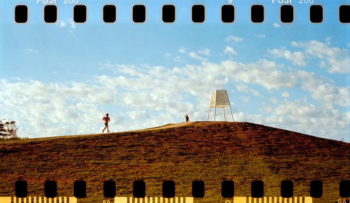 A man runs along a path on a grassy hill with a white structure and another person in the distance under a cloudy sky.