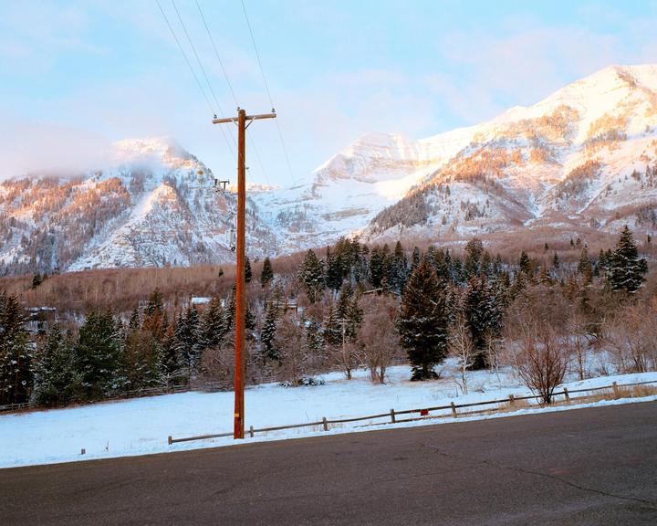 A snow-covered mountain range with pine trees and a utility pole in the foreground is illuminated by the setting sun.