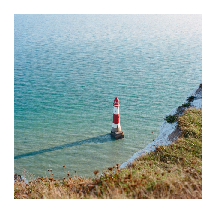 A red and white striped lighthouse stands in the turquoise sea, casting a long shadow on the water, with a grassy cliff edge in the foreground.