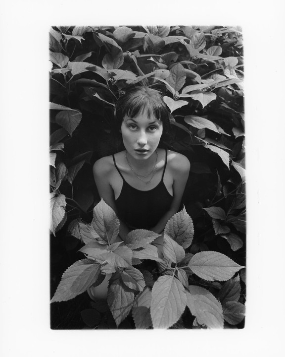 A woman with short hair and a necklace is surrounded by large leaves.