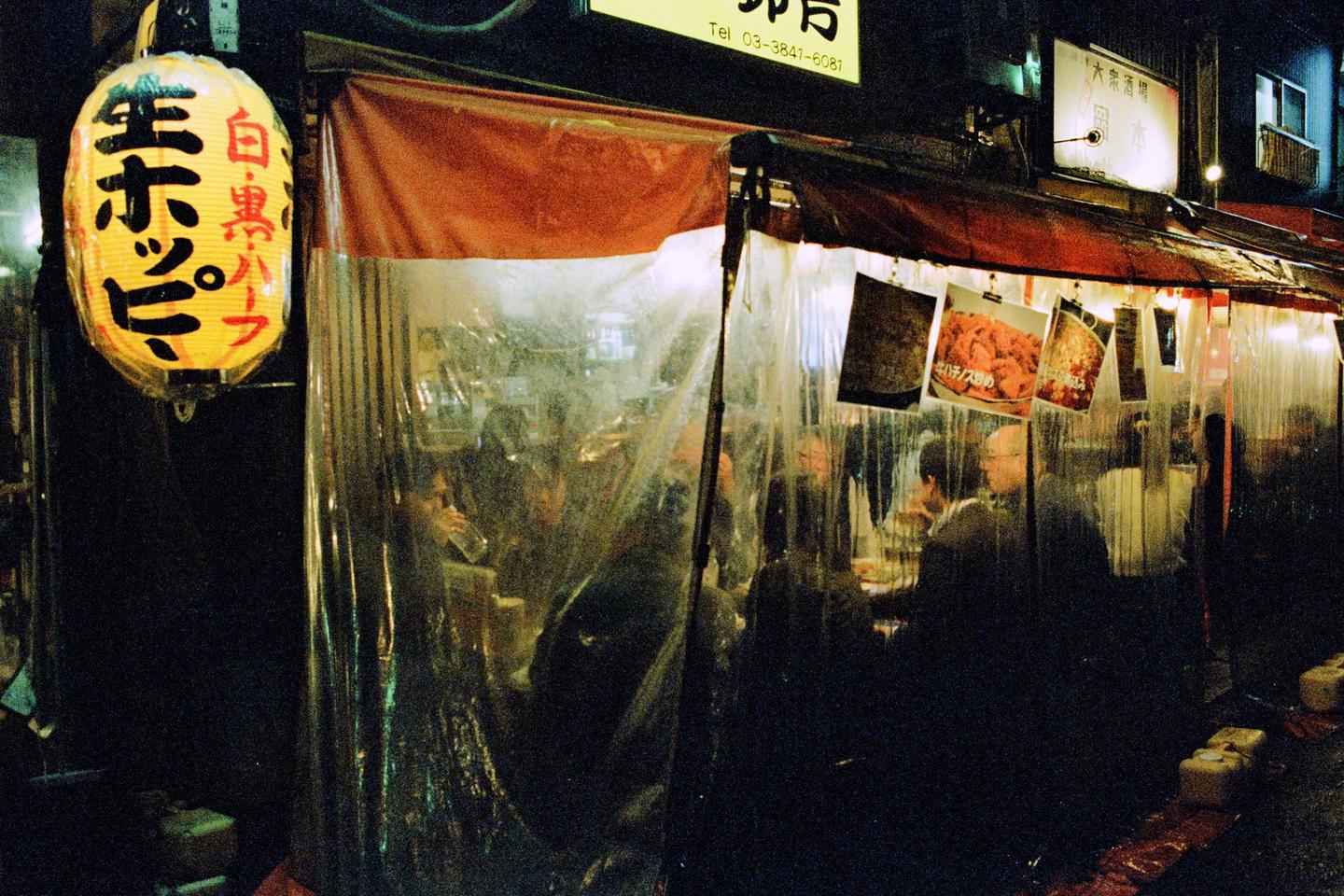 A group of people are seated at tables inside a restaurant with plastic sheeting for walls, next to a street with a yellow lantern.