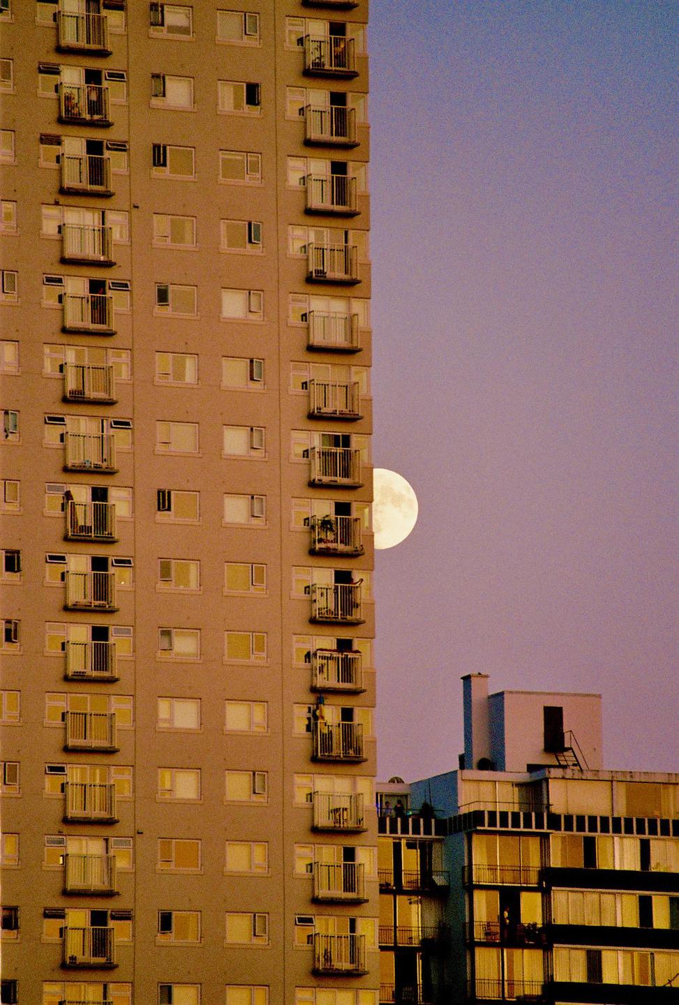A full moon is visible behind a tall apartment building with many balconies.