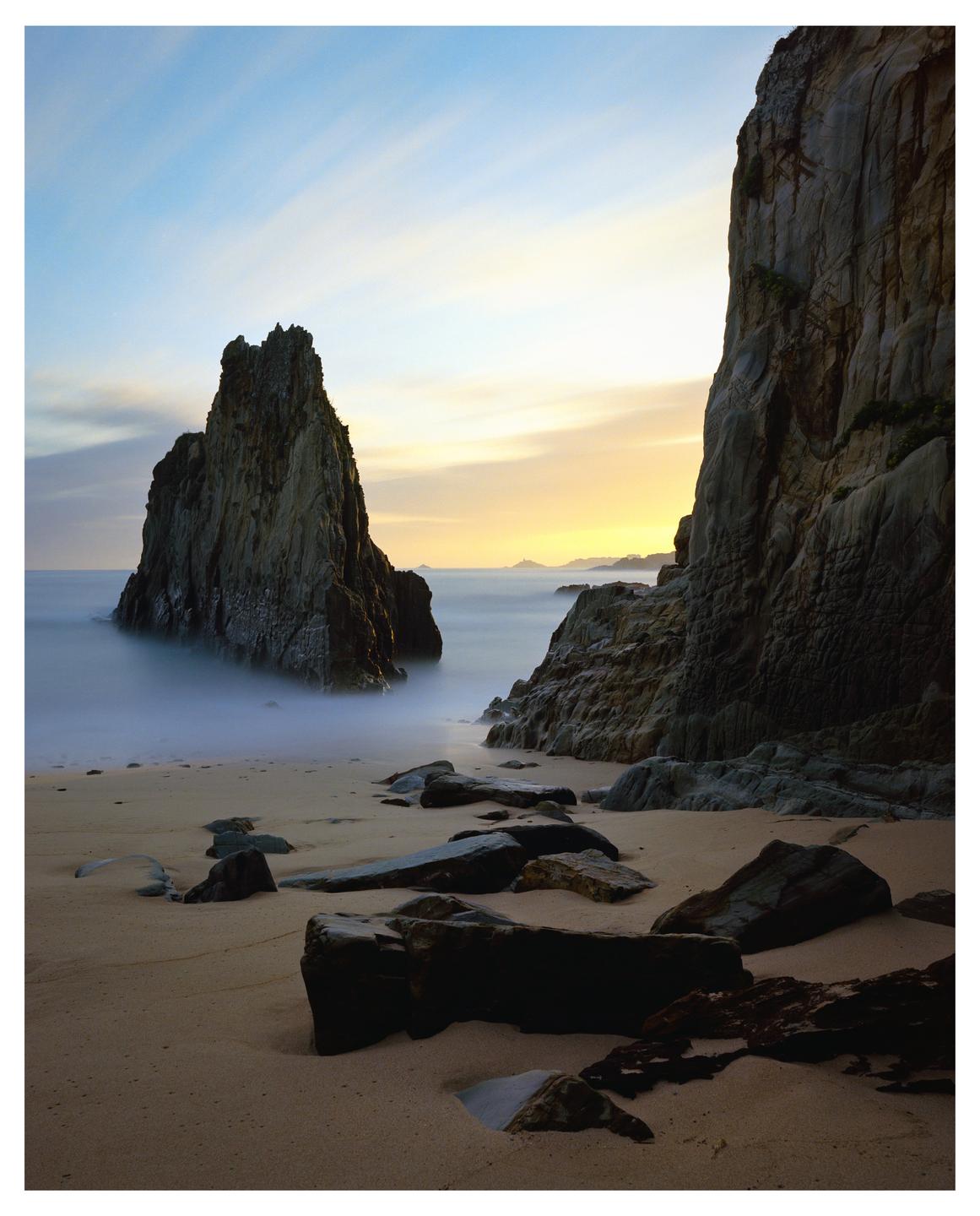 Two large rock formations stand on a sandy beach with the ocean and a colorful sky in the background.