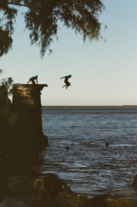 A person jumps from a stone wall into the ocean while another person watches from the wall.