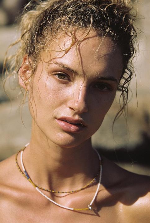 A woman with wet, curly hair and two beaded necklaces looks directly at the viewer.