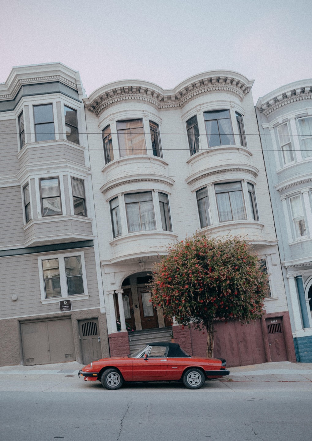 A red convertible car is parked on a street in front of a white building with bay windows and a flowering tree.