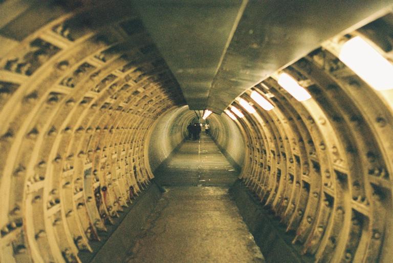 A long, curved tunnel with exposed metal ribs and lights on the ceiling leads to a group of people in the distance.