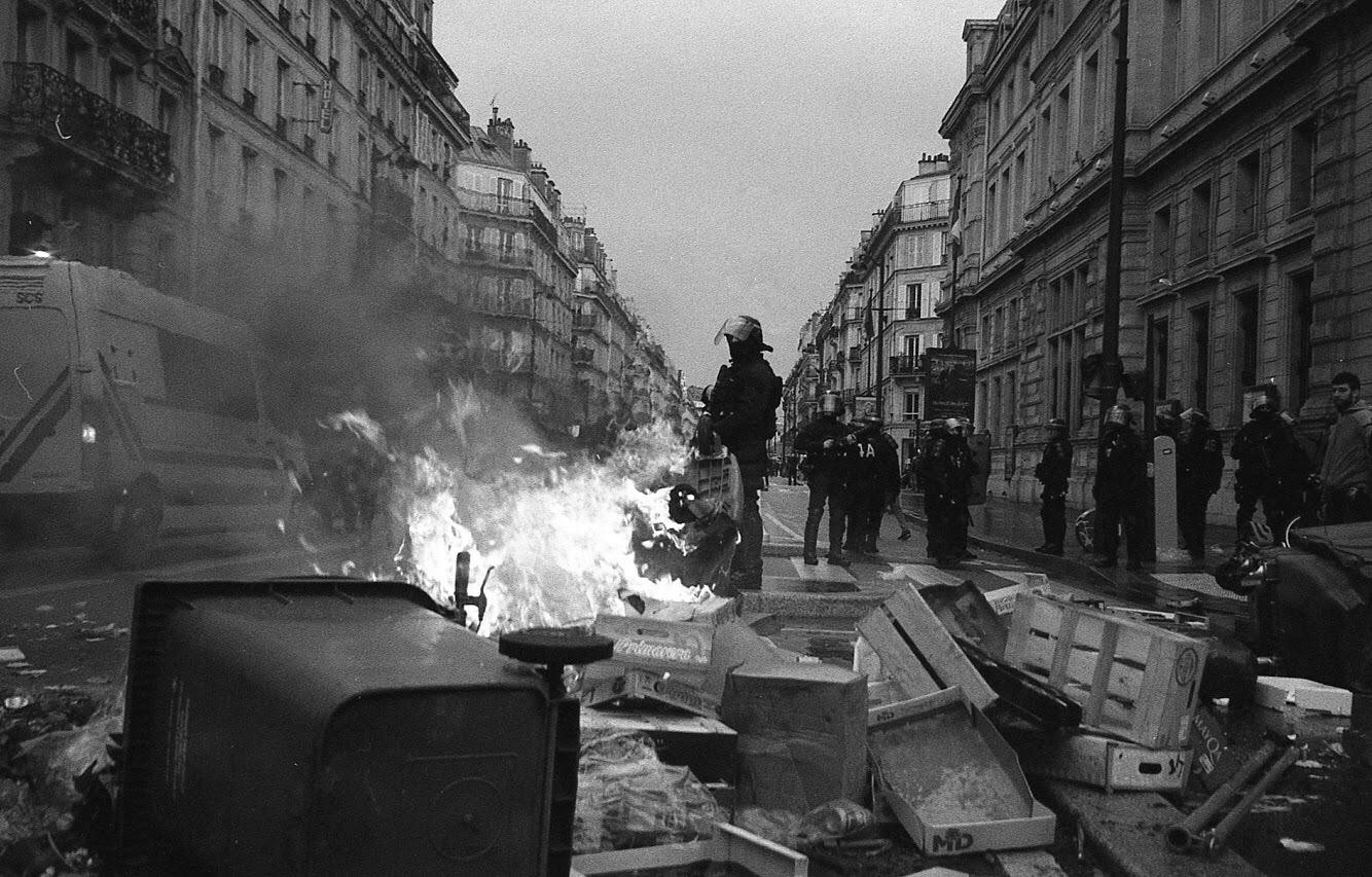 A street in Paris is filled with smoke and fire from burning debris, with police officers standing amidst the chaos and buildings lining both sides of the road.