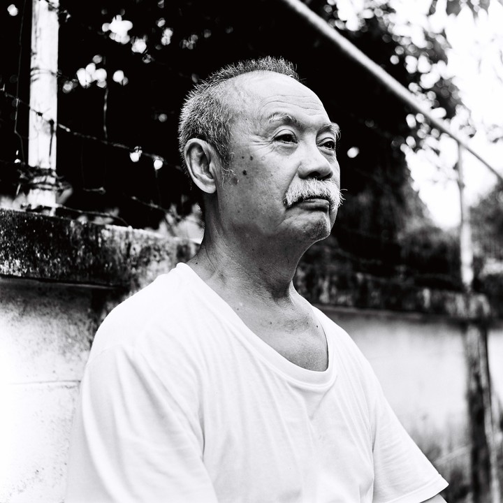 A man with a mustache looks to the right of the frame, with a wall and foliage in the background.