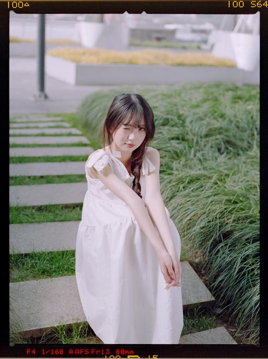 A woman in a white dress winks at the camera while sitting on a stone path surrounded by grass.