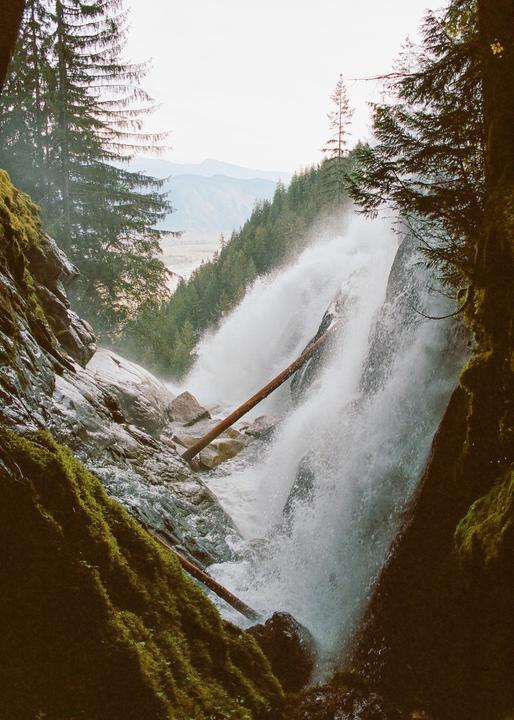 A powerful waterfall cascades over rocks and fallen logs in a forest with mountains in the distance.