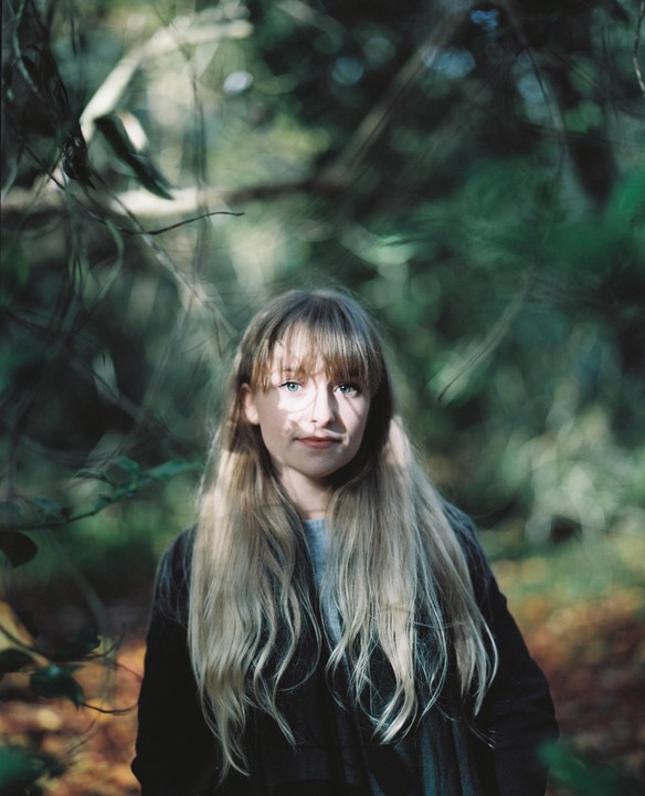 A girl with long blonde hair stands in a forest with dappled sunlight on her face.