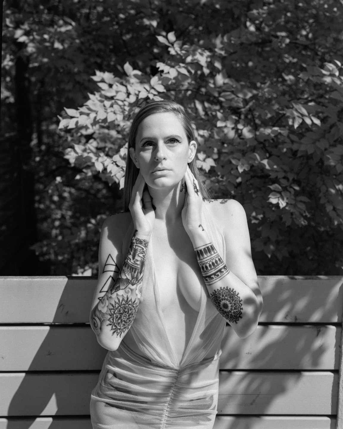 A woman with tattoos on her arms poses with her hands around her neck, in front of a wooden fence and foliage.