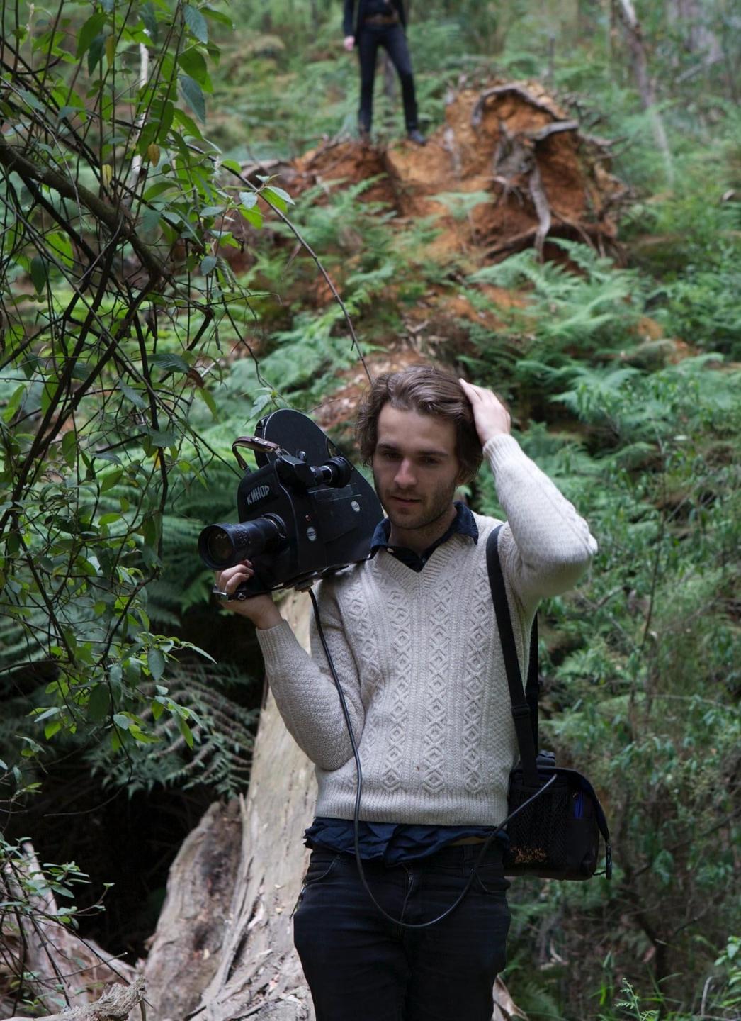 A man holds a large camera in a forest with another person standing on a tree root in the background.