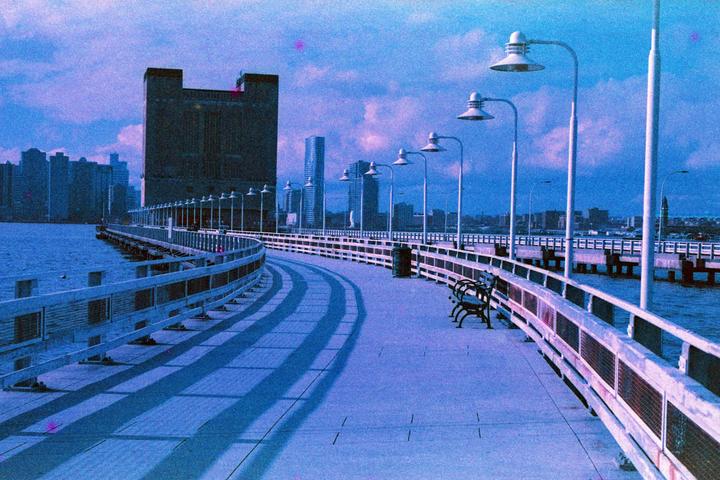 A long pier with streetlights and benches curves along a body of water, with a city skyline in the background.